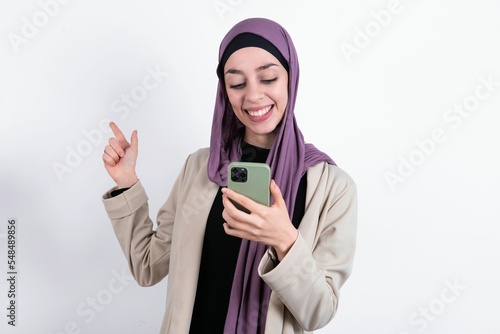 Smiling young beautiful muslim woman wearing hijab and jacket over white background pointing finger at blank space holding phone in one hand © Jihan