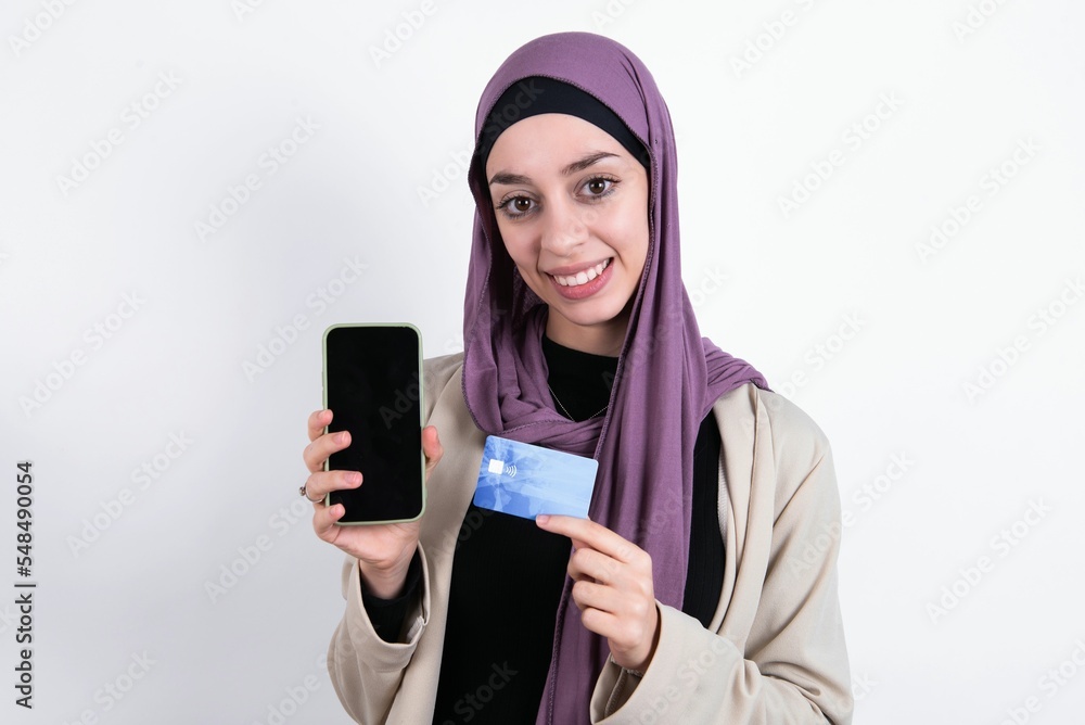 Photo of adorable young beautiful muslim woman wearing hijab and jacket over white background holding credit card and Smartphone. Reserved for online purchases Photo of adorable young beautiful muslim woman wearing hijab and jacket over white background holding credit card and Smartphone. Reserved for online purchases
