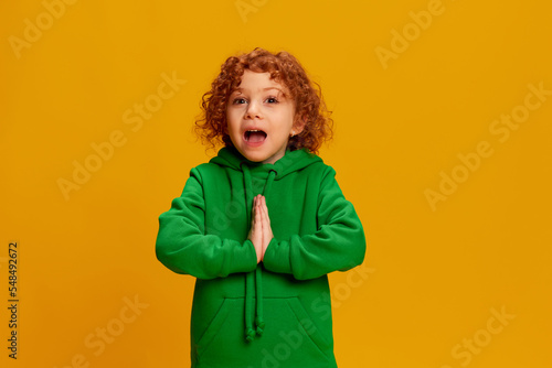 Portrait of cute little girl, child with curly red hair emotionally posing isolated over yellow background