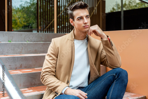 Young guy with modern hairstyle with coat, scarf, blue jeans and white sweeter sitting outdoors