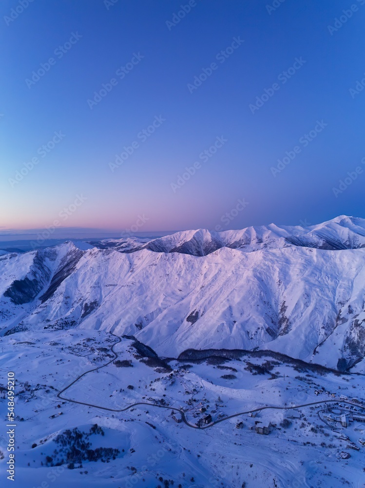 Fototapeta premium Aerial of snowy mountain range on winter sunrise at ski resort. Mountains valley and village with switchbacks road at sunset. Caucasus peaks skyline in the dusk. City lights at night.