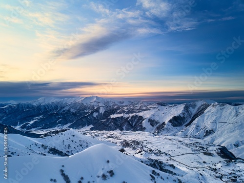 Wide aerial panorama of snowy mountain ridge at ski resort village on sunrise. Stunning mountains range covered with snow powder on ski resort at sunset. Caucasus mountain peaks skyline on sunset.