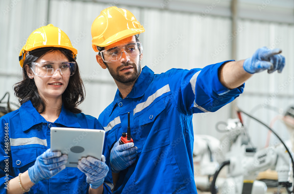 Industrial engineer manager and woman colleague standing using digital tablet computer to control robot automated machines in smart factory. Manufacturing industry technology innovation workshop. Industrial engineer manager and woman colleague standing using digital tablet computer to control robot automated machines in smart factory. Manufacturing industry technology innovation workshop.