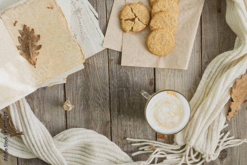 On the wooden table there is a mug of hot coffee with foam, cookies, a book, a branch of mountain ash and a warm scarf. The concept of warmth, comfort