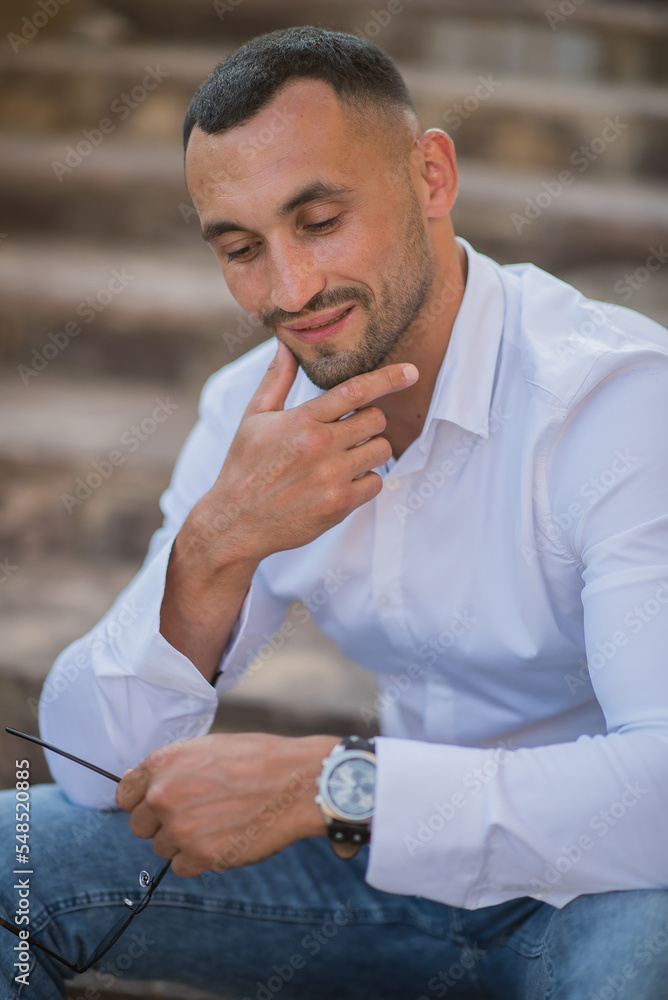 Portrait of a handsome confident stylish man. Sexy modern man wearing a white shirt. Fashionable man posing against the backdrop of a street in the city at sunset, holding sunglasses in his hands Portrait of a handsome confident stylish man. Sexy modern man wearing a white shirt. Fashionable man posing against the backdrop of a street in the city at sunset, holding sunglasses in his hands