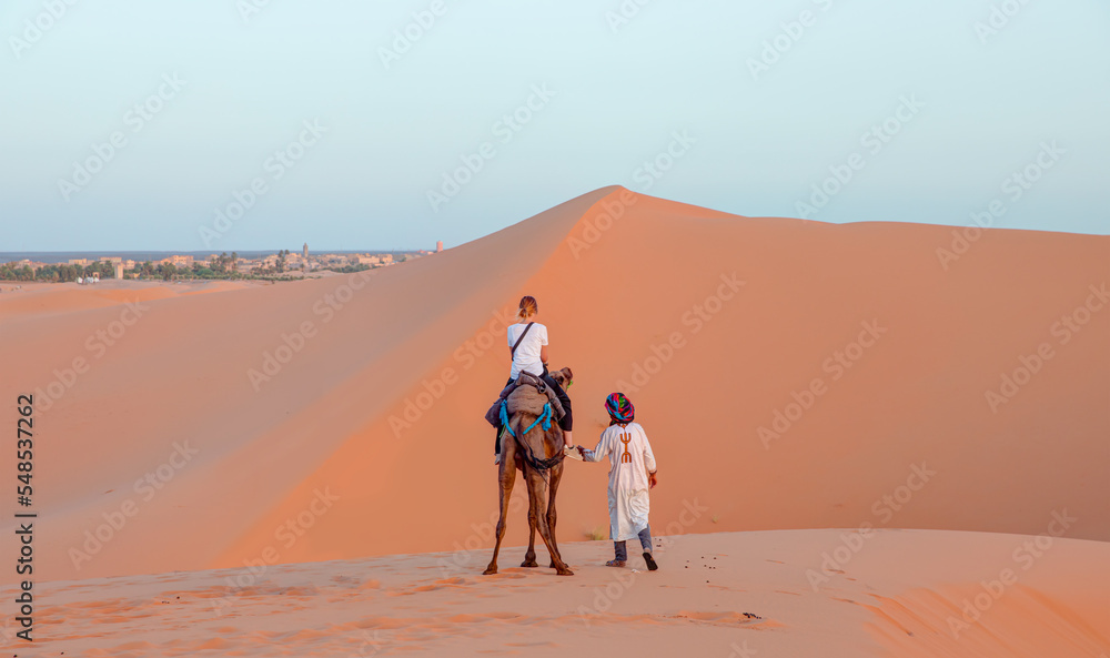 A woman riding a camel across the thin sand dunes of the in Western Sahara Desert, Morocco, Africa A woman riding a camel across the thin sand dunes of the in Western Sahara Desert, Morocco, Africa