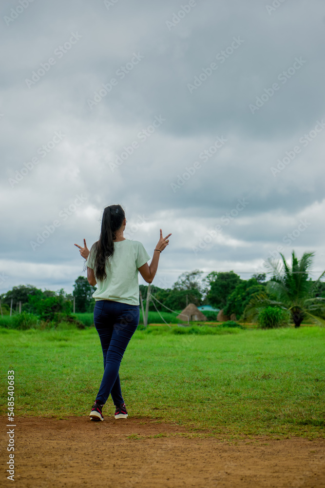 Young attractive model standing in beautiful green grass field, showing peace gesture with both hands from behind.