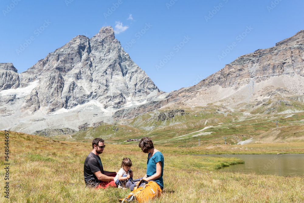 Toddler drinking water from a hydration bag while is resting with her parents on a green meadow under the Matterhorn at italian alps.