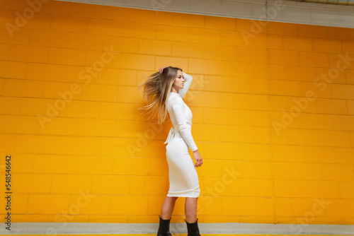 Fashionable beautiful girl with flowing hair in a stylish white dress walks near a yellow brick wall. Urban pretty woman in parking lot indoors