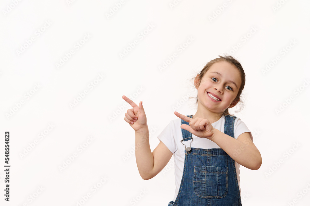 Adorable delightful Caucasian little child, a cute baby girl wearing a blue denim overalls, smiling a toothy smile, pointing at a copy space for promotional text, isolated over white background. Adorable delightful Caucasian little child, a cute baby girl wearing a blue denim overalls, smiling a toothy smile, pointing at a copy space for promotional text, isolated over white background.