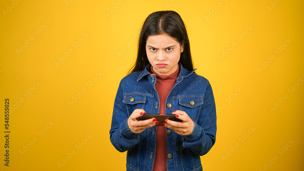Sad or Angry modern gamer girl using mobile phone, Aggressive or shocked young woman playing video game on smartphone, lost the game, standing isolated over yellow background Sad or Angry modern gamer girl using mobile phone, Aggressive or shocked young woman playing video game on smartphone, lost the game, standing isolated over yellow background