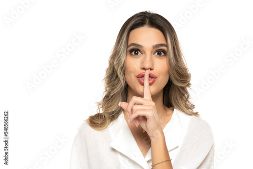 cute woman with long wavy hair holding her finger on her mouth as she was keeping a secret on a white studio background