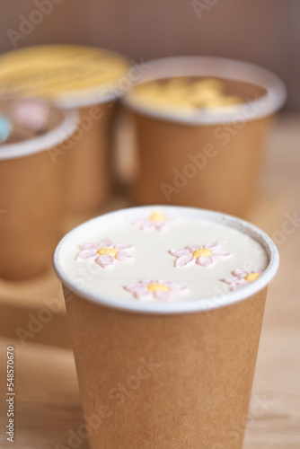 a paper cup with cake to go with cream drawing flowers on a table and three homemade mini chocolate cake in coffee cups in a box on background, confectionery trend concept. Vertical, copy space, Bokeh