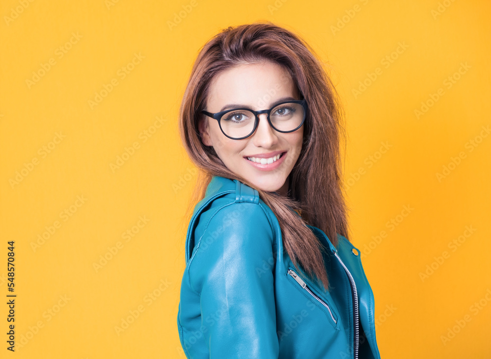 Face of nice girl keeps chin on shoulder, wearing blue leather jacket, isolated on yellow background. Closeup portrait of a cheerful woman