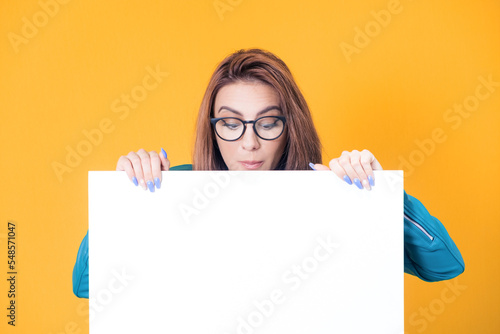 Close-up woman behind white board and looking down to empty space, wearing blue leather jacket, isolated on yellow background. Pretty girl showing blank space for text near her head