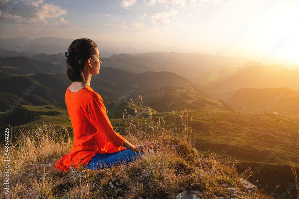 young caucasian woman meditates in the lotus position in the grass in the mountains near the cliff in the setting sun