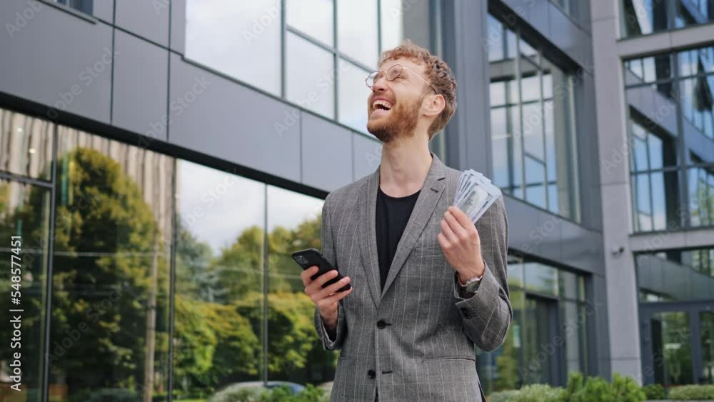 Young excited joyful caucasian man wearing fashion suit and glasses using mobile cell phone hold win fan of cash money in dollar banknotes standing near stock market celebrate good news success. Young excited joyful caucasian man wearing fashion suit and glasses using mobile cell phone hold win fan of cash money in dollar banknotes standing near stock market celebrate good news success.