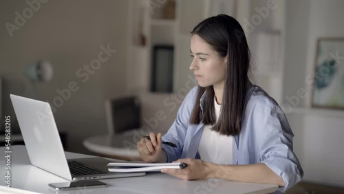 Young female project manager making online video chat meeting with clients and making notes in her notebook. Remote conference with team and colleagues via webcam on laptop working from home photo