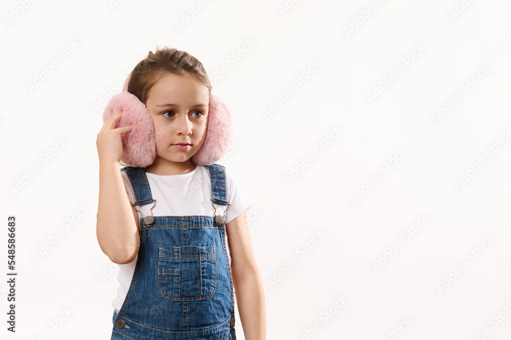 Beautiful Caucasian little child girl wearing blue denim overalls, cute pink plush headphones - winter earmuffs, ear warmers, isolated on white background. Protect your ears from cold. Copy space Beautiful Caucasian little child girl wearing blue denim overalls, cute pink plush headphones - winter earmuffs, ear warmers, isolated on white background. Protect your ears from cold. Copy space
