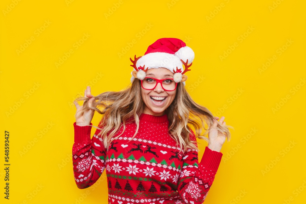 A young happy smiling blonde woman in a Christmas red deer sweater, glasses with deer antlers and a Santa Claus hat holds strands of hair in her hands isolated on a solid color yellow background A young happy smiling blonde woman in a Christmas red deer sweater, glasses with deer antlers and a Santa Claus hat holds strands of hair in her hands isolated on a solid color yellow background