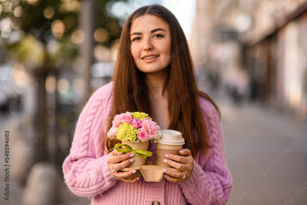 Fototapeta premium handsome woman gently holds flower arrangement in small pot and paper cup of coffee in her hands.