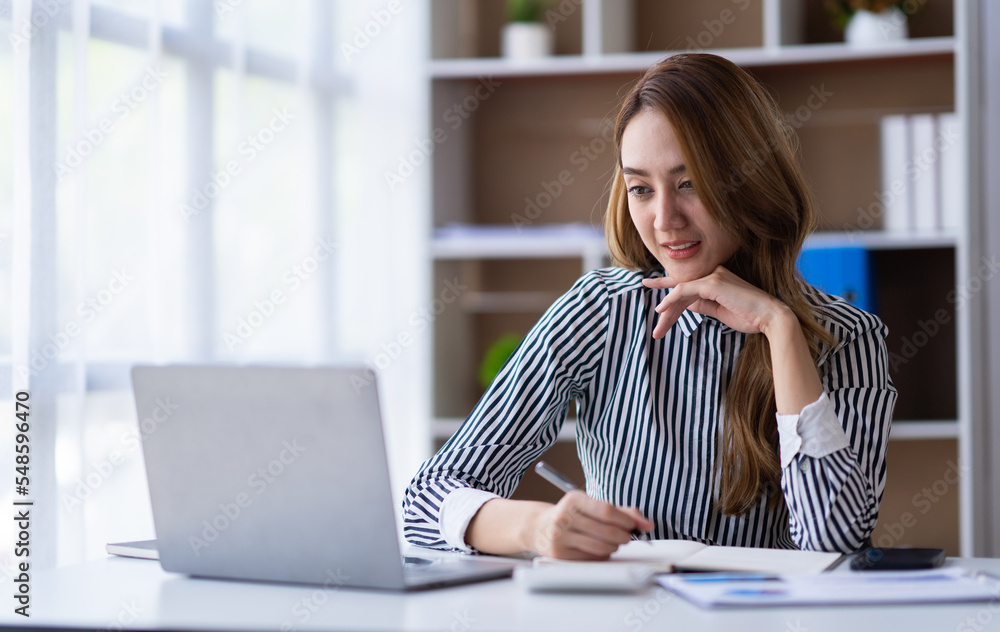 Portrait of a cheerful Asian businesswoman using a laptop and writing on a charger to keep notes as a base Portrait of a cheerful Asian businesswoman using a laptop and writing on a charger to keep notes as a base