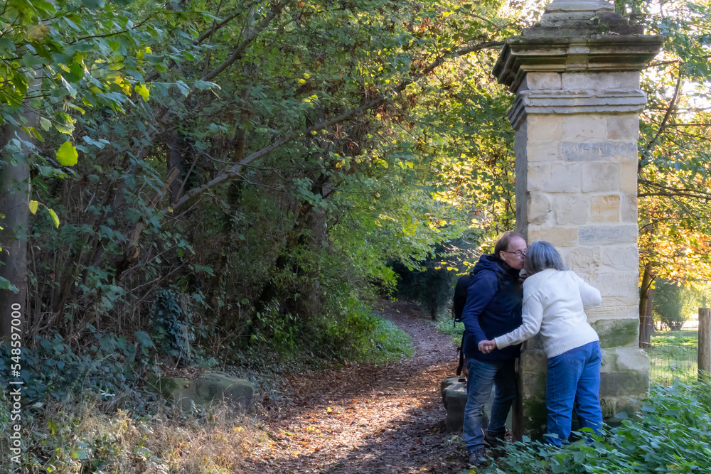 Senior couple kissing on old pillar at entrance to Vliekerbos nature reserve, trail and yellowish-green foliage in background, Landgoed Vliek castle gardens, autumn day in South Limburg, Netherlands Senior couple kissing on old pillar at entrance to Vliekerbos nature reserve, trail and yellowish-green foliage in background, Landgoed Vliek castle gardens, autumn day in South Limburg, Netherlands