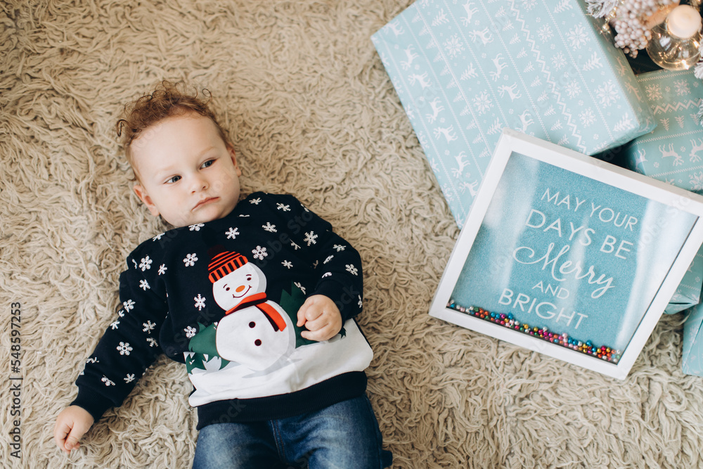 Naklejka premium A little curly boy in a traditional Christmas sweater spends time at home near the Christmas tree