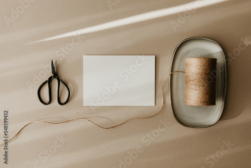 Stylish flat lay with a white blank paper greeting card, a black metal scissors, and a roll of twine string. Top view, copy space image. Creative message concept on beige background. Autumn colors.
