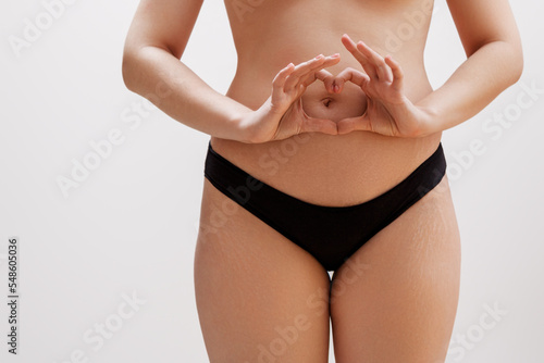 Self loving woman making a heart shape on her stomach in a studio. Closeup of an anonymous woman standing in black underwear. Young woman embracing her natural body against a studio background.