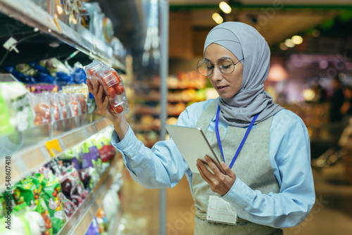 A Muslim woman in a hijab, a supermarket worker checks products on shelves and in refrigerators, a woman with a tablet and an apron uses a tablet to review products.