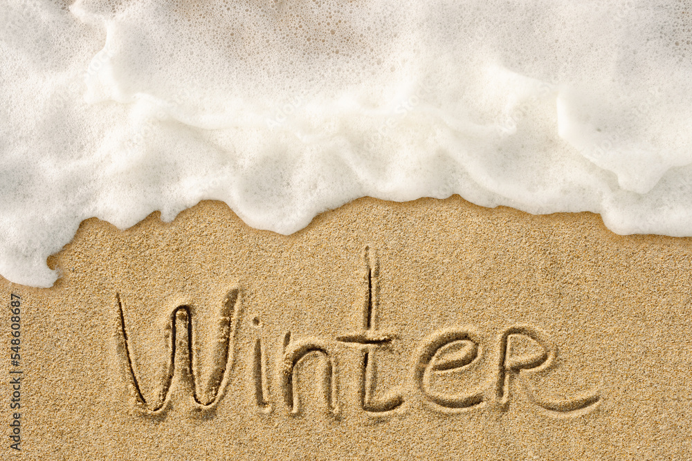 Fototapeta premium Winter word handwritten lettering on the sandy beach with white ocean wave background, seasonal concept photo