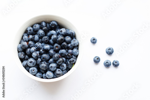 A white bowl full of blueberries stands on a white table and five berries lie nearby. View from above. Soft focus.