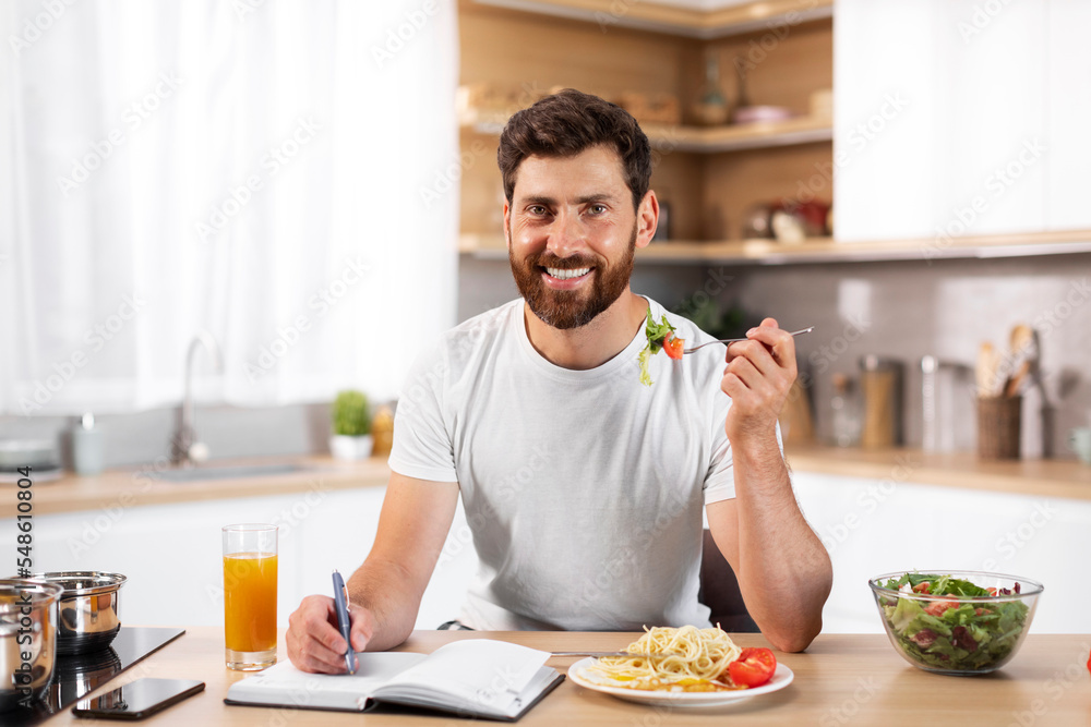 Smiling middle aged european bearded man making notes, planning business and eating pasta