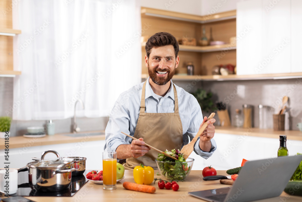 Smiling adult european bearded male chef in apron preparing salad in modern kitchen interior Smiling adult european bearded male chef in apron preparing salad in modern kitchen interior