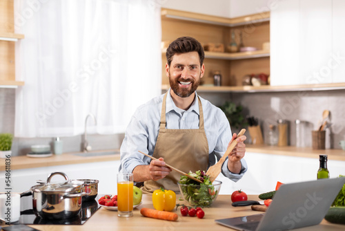 Smiling adult european bearded male chef in apron preparing salad in modern kitchen interior
