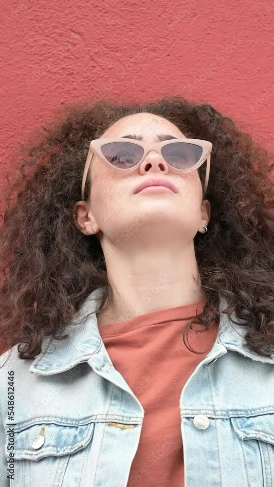 Vertical video.Portrait of a woman with curly hair and sunglasses. Leaning against a red concrete wall. Young woman with afro hair looking upwards. Vertical video.Portrait of a woman with curly hair and sunglasses. Leaning against a red concrete wall. Young woman with afro hair looking upwards.