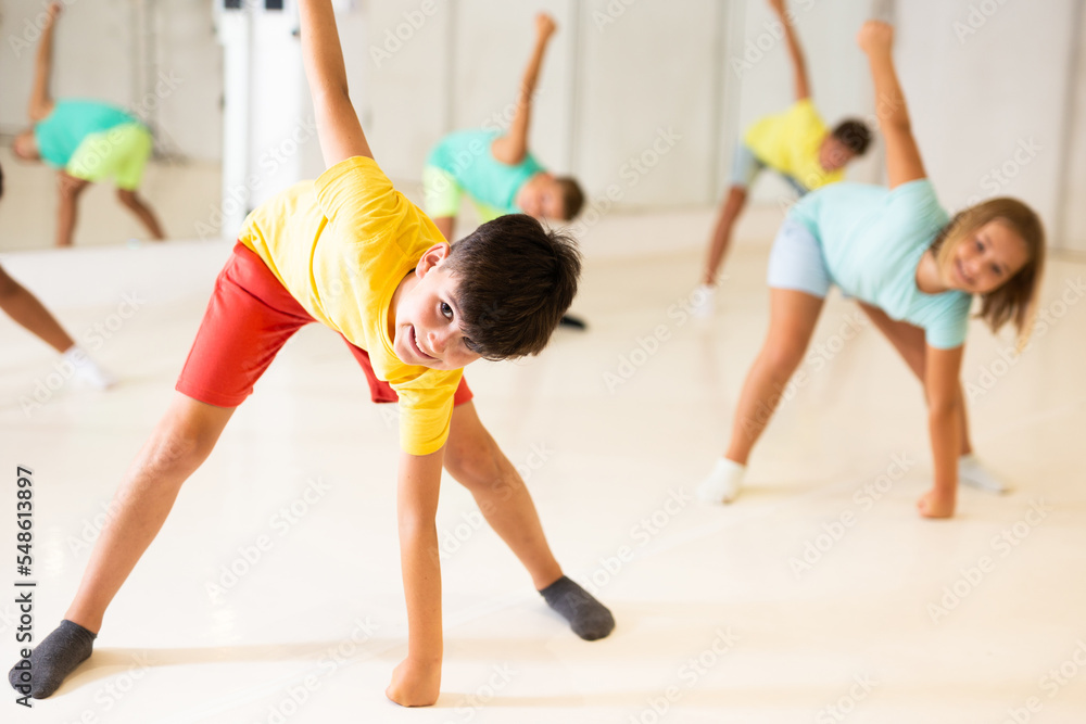 Smiling preteen boy doing stretching workout with group of children before dance training in choreography class.