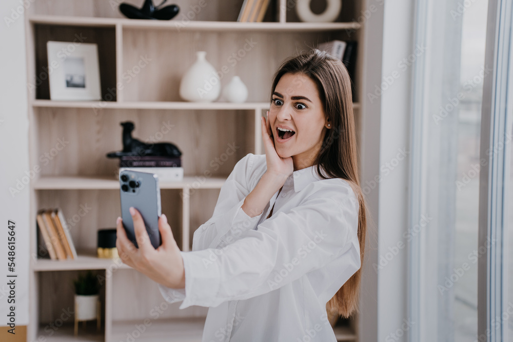 Shocked successful brunette woman at home in white shirt makes video call with wide opened eyes and mouth against blurry interior. Excited pretty girl receives good news, she got a job. Amazed blogger Shocked successful brunette woman at home in white shirt makes video call with wide opened eyes and mouth against blurry interior. Excited pretty girl receives good news, she got a job. Amazed blogger