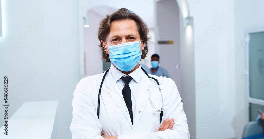 Close up portrait of Caucasian handsome male doctor wearing medical mask standing in clinic at reception and looking at camera. Mixed-races healthcare workers working on background. Covid 19 concept Close up portrait of Caucasian handsome male doctor wearing medical mask standing in clinic at reception and looking at camera. Mixed-races healthcare workers working on background. Covid 19 concept