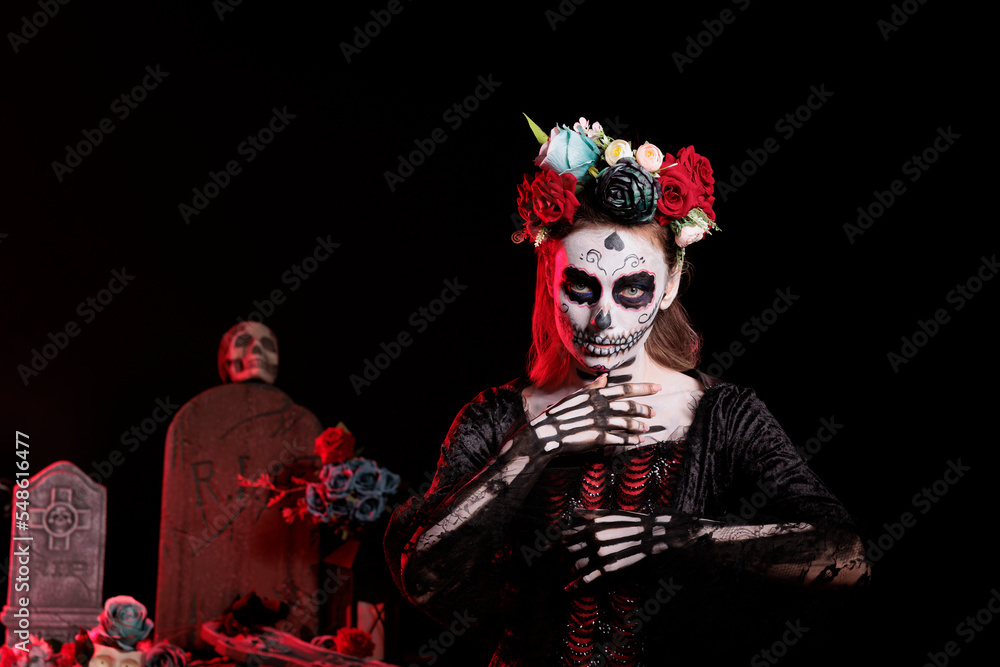Fototapeta premium Horror lady goddess of death costume with body art, wearing flowers crown to celebrate dios de los muertos tranditional mexican holiday. Woman with santa muerte skull make up in studio.