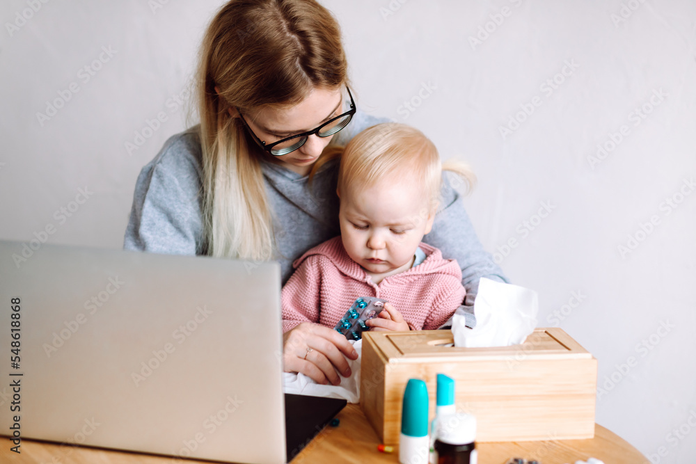 Portrait of young mother sitting at table near box, holding little baby, looking at pills blister at home. Sick time. Portrait of young mother sitting at table near box, holding little baby, looking at pills blister at home. Sick time.