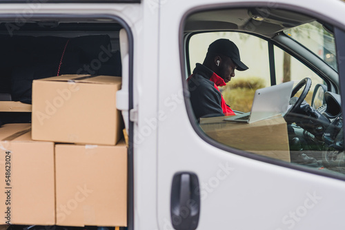 Modern courrier. Online shopping package delivery concept. White delivery van with opened side doors revealing many undelivered packages. Adult Black deliveryman in dark-colored hat and red jacket photo