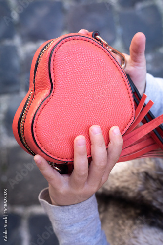 Baby holds a small heart shaped red bag. Grey background. Accessories and love contest. Little lady. Saint Valentines day present. Expressing love and tender feelings. photo