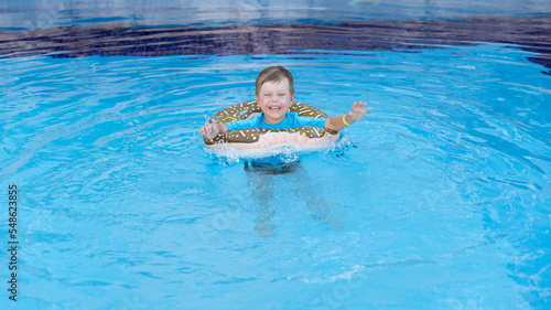 A little happy boy bathes in a doughnut lifebuoy in clear water in swimming pool in sunny weather