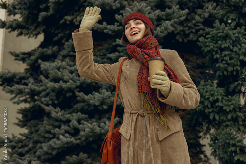 Happy woman in outerwear with cup of coffee to go waving hand and greeting friend in park in winter 