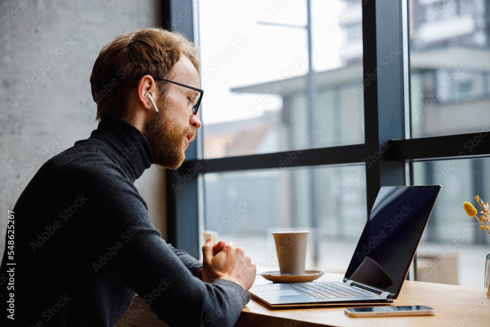 A young red-haired guy, a programmer or businessman in glasses, is sitting in a stylish cafe at a laptop and is talking through a webcam. Freelancer works remotely. Online communication. A young red-haired guy, a programmer or businessman in glasses, is sitting in a stylish cafe at a laptop and is talking through a webcam. Freelancer works remotely. Online communication.