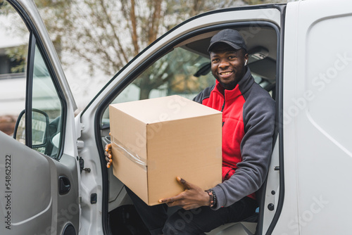 Friendly smiling Black man in his mid 20 sitting on driver's seat with opened door and holding cardboard package. Looking at camera. Positive vibes. High quality photo