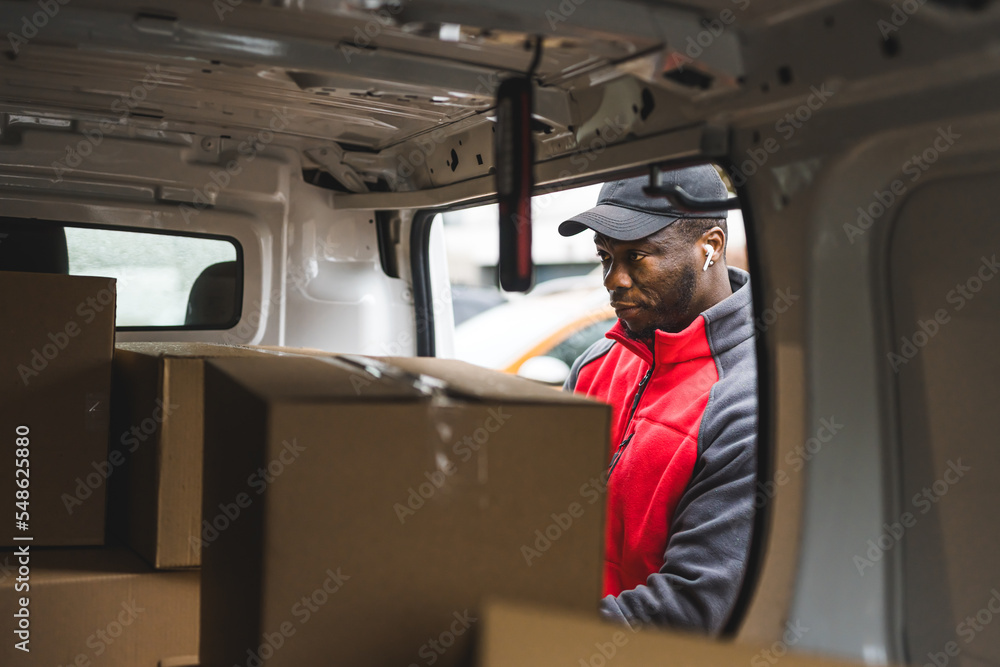 Young adult black delivery man wearing red uniform pullover looking inside van full of carboard box parcels. Horizontal shot. High quality photo Young adult black delivery man wearing red uniform pullover looking inside van full of carboard box parcels. Horizontal shot. High quality photo