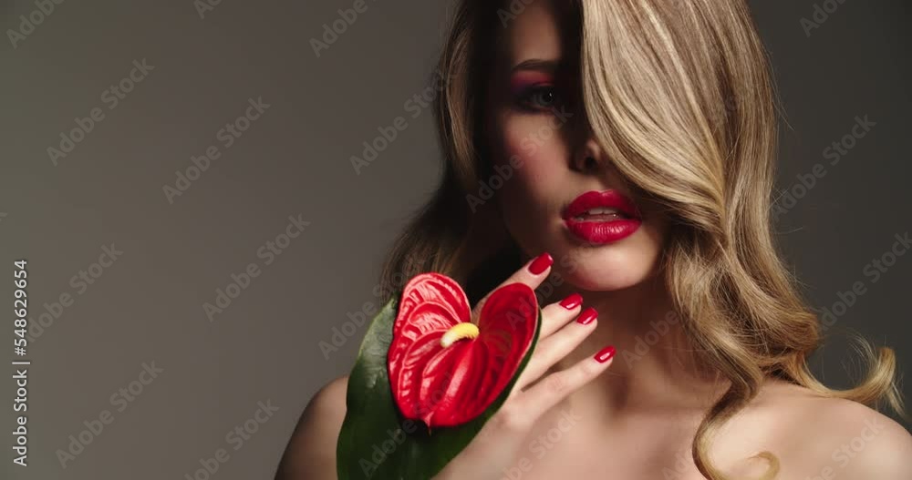 Sexy blonde woman with red lips holds a red flower. Fashion model with a long wavy hair posing at studio. Closeup face of a pretty young woman with bright makeup. Slow motion. Sexy blonde woman with red lips holds a red flower. Fashion model with a long wavy hair posing at studio. Closeup face of a pretty young woman with bright makeup. Slow motion.
